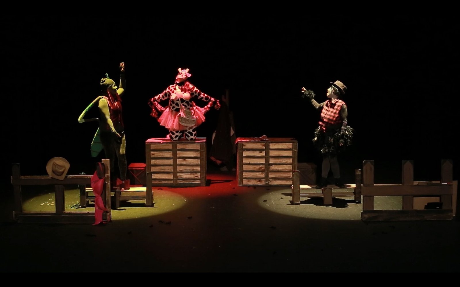 Escena teatral con tres personajes en vestuario colorido sobre fondo negro